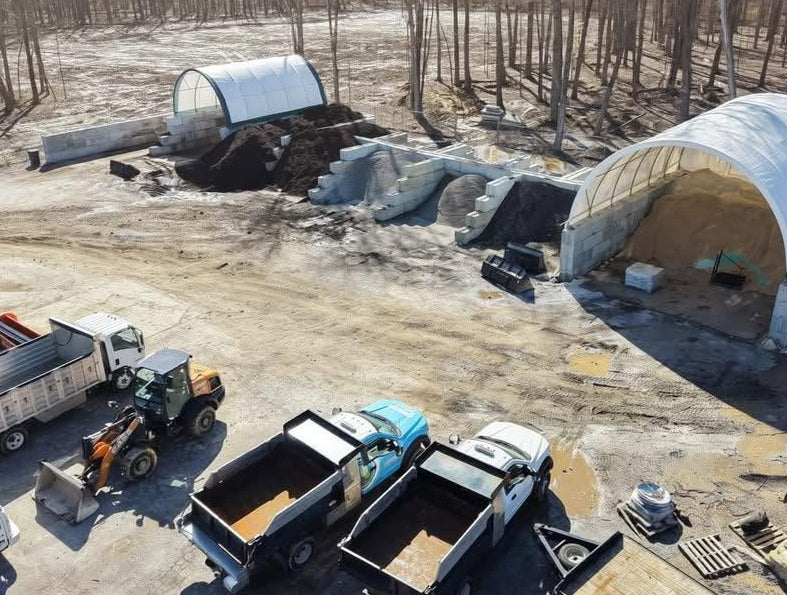 An aerial view of a construction or landscaping site with two large white arched sheds, several trucks, a loader, and bare trees in the background.