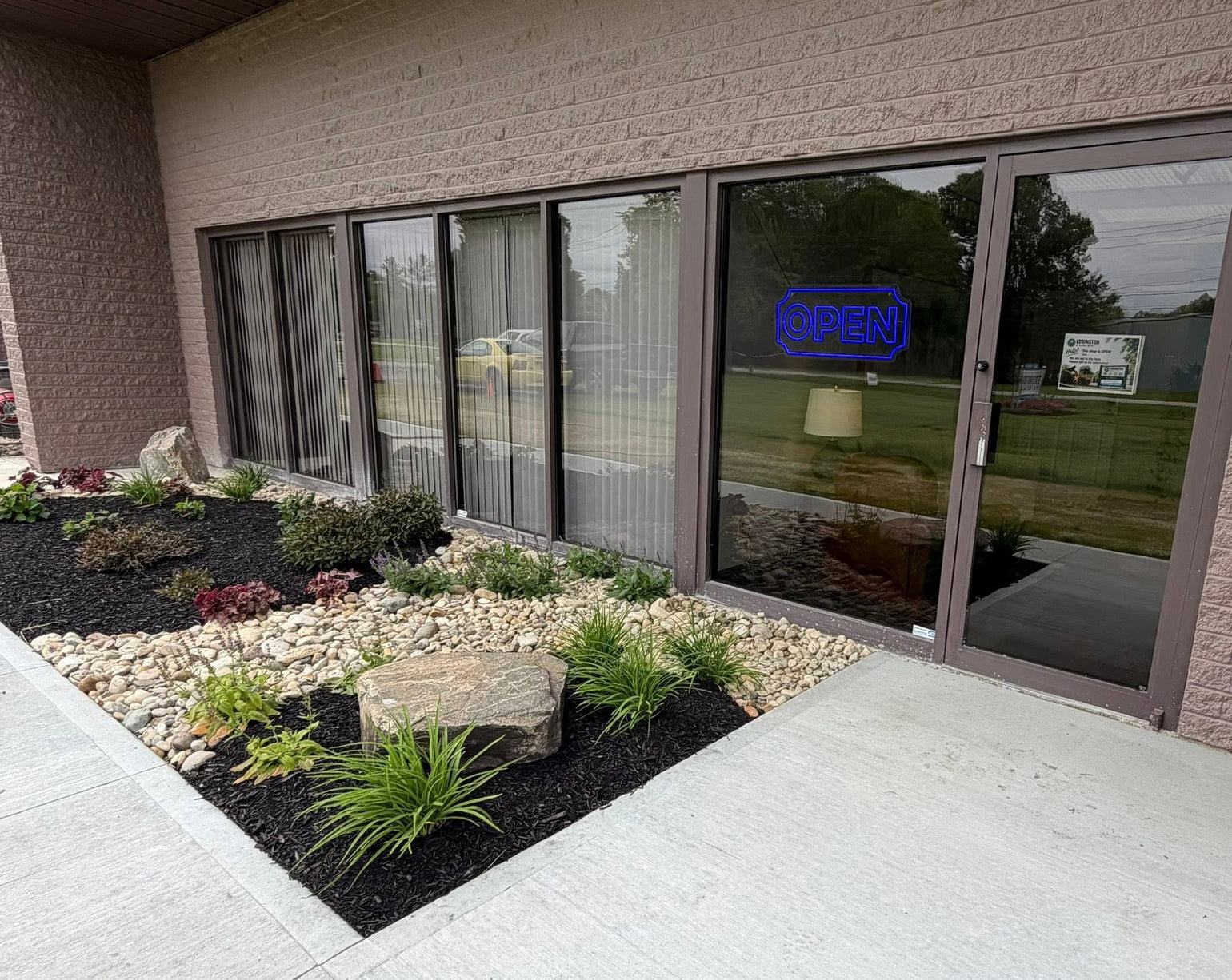 A landscaped building entrance with a walkway, black mulch beds, white gravel, small plants, and a stone feature in front of large glass doors and windows. A blue neon-style OPEN sign is visible on one door.